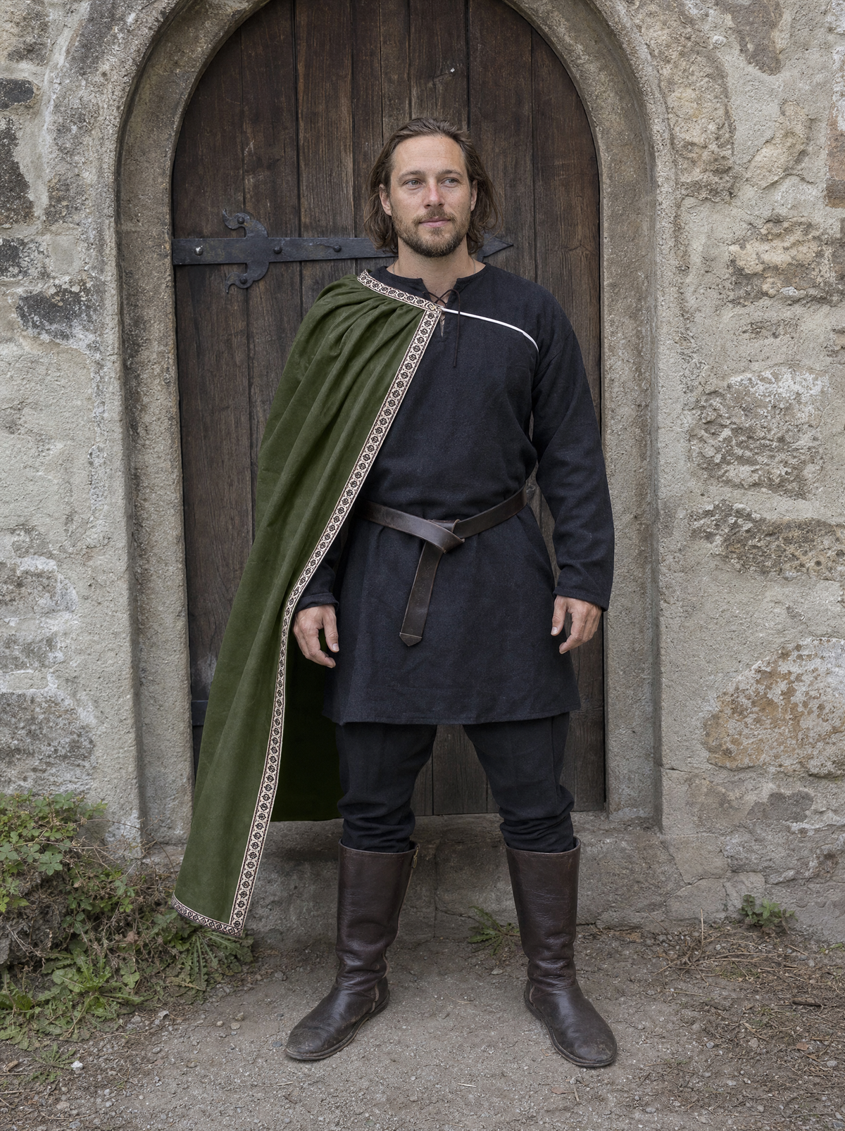Man in a dark tunic with a green velvet cape over one shoulder by an arched stone doorway