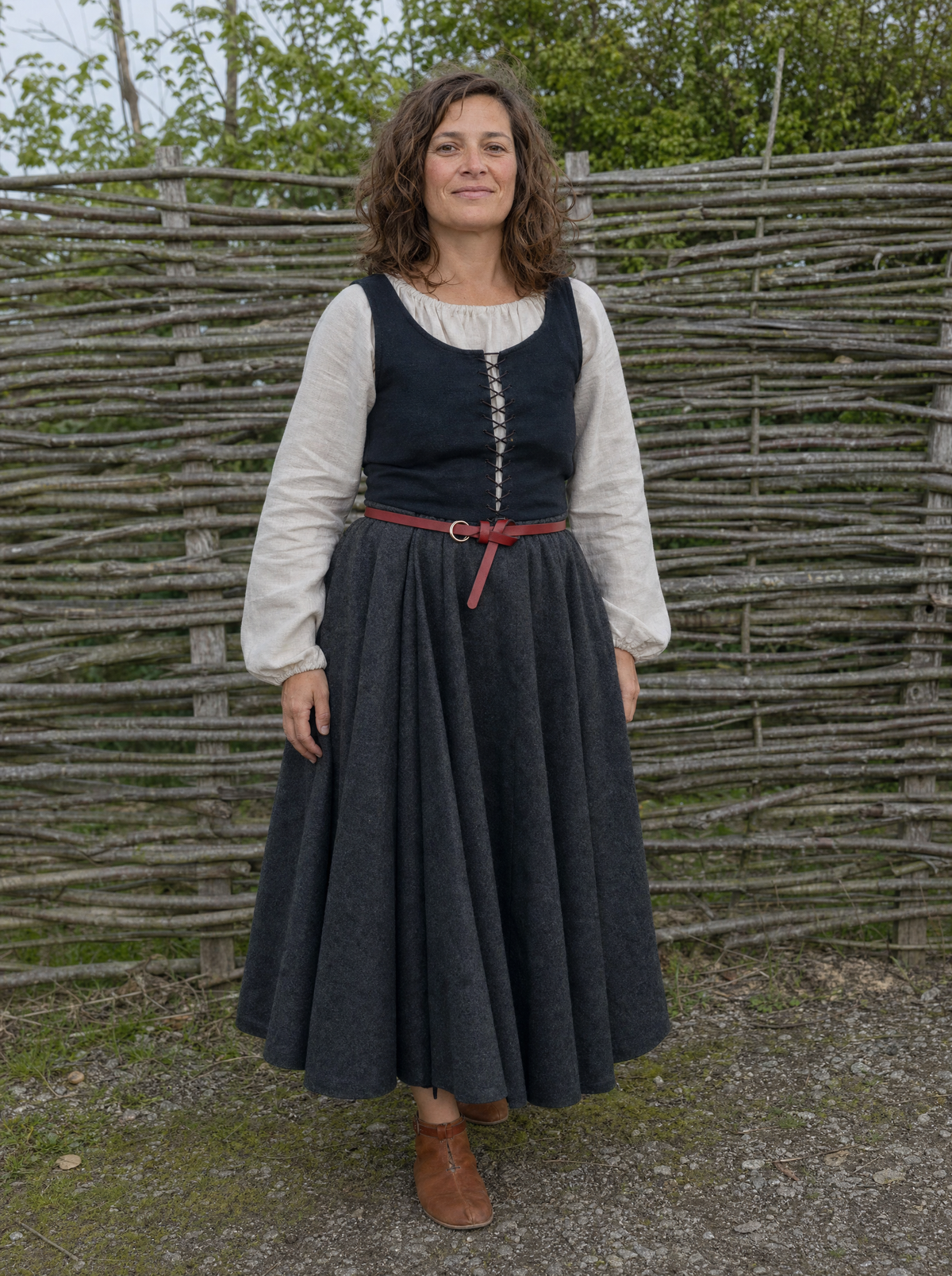 Woman in a white blouse, black laced vest and long skirt with a thin red belt by a wattle fence
