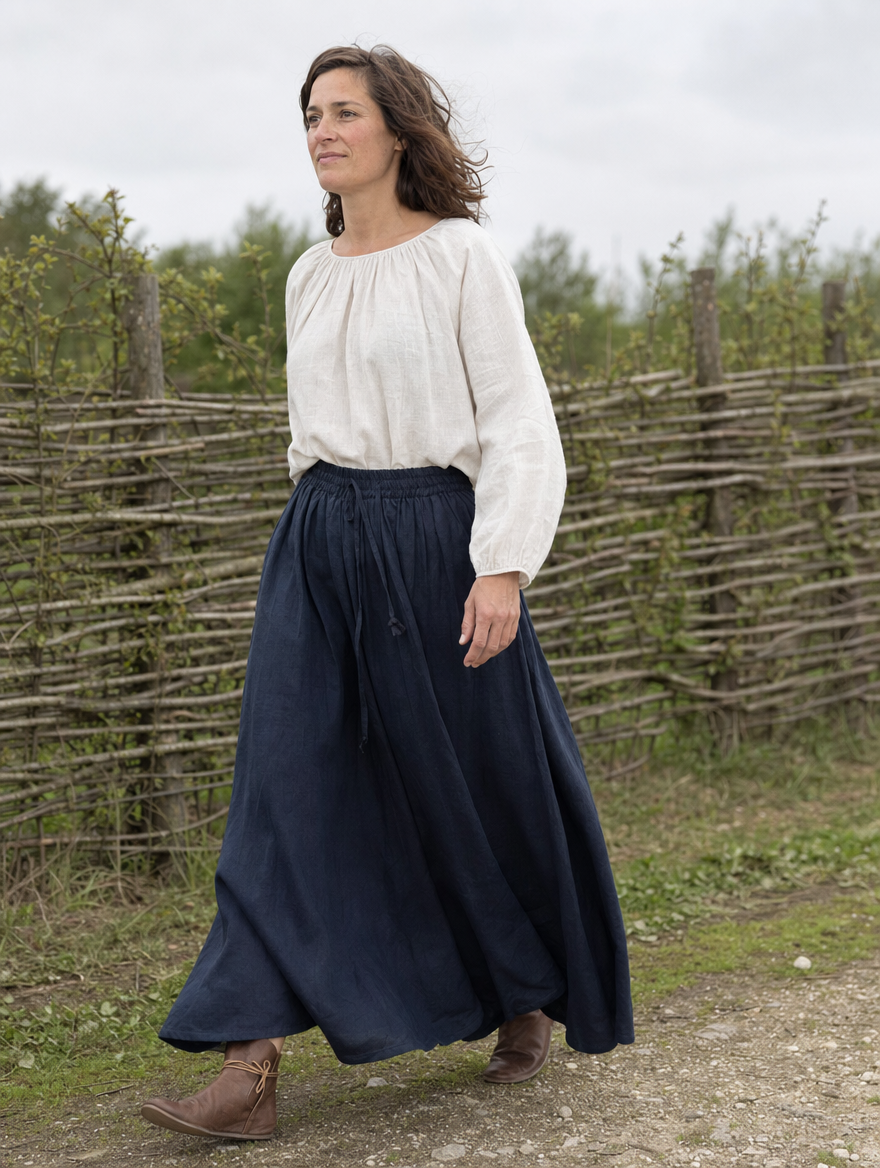 Woman in a loose white blouse and a long navy skirt walking beside a wattle fence